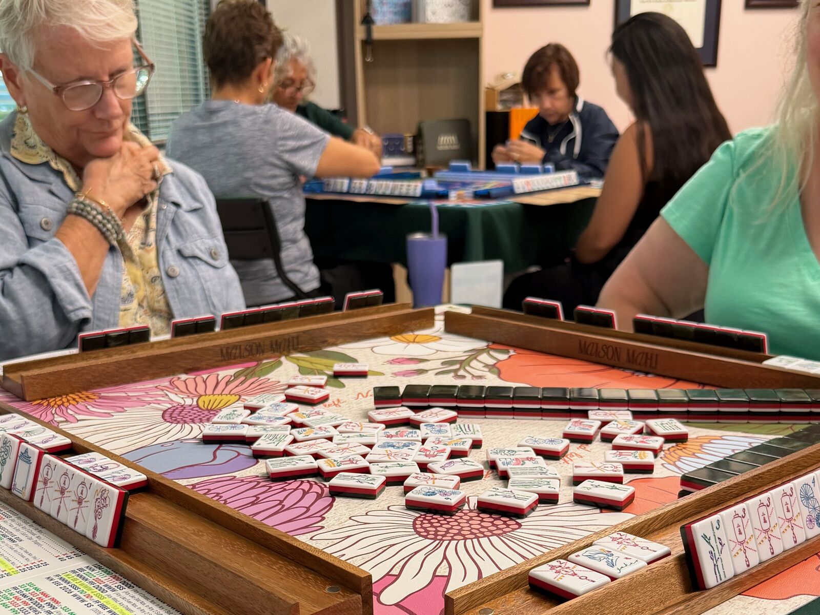 A floral-print Mahjong mat with tiles and wooden racks, with players at other tables in the background