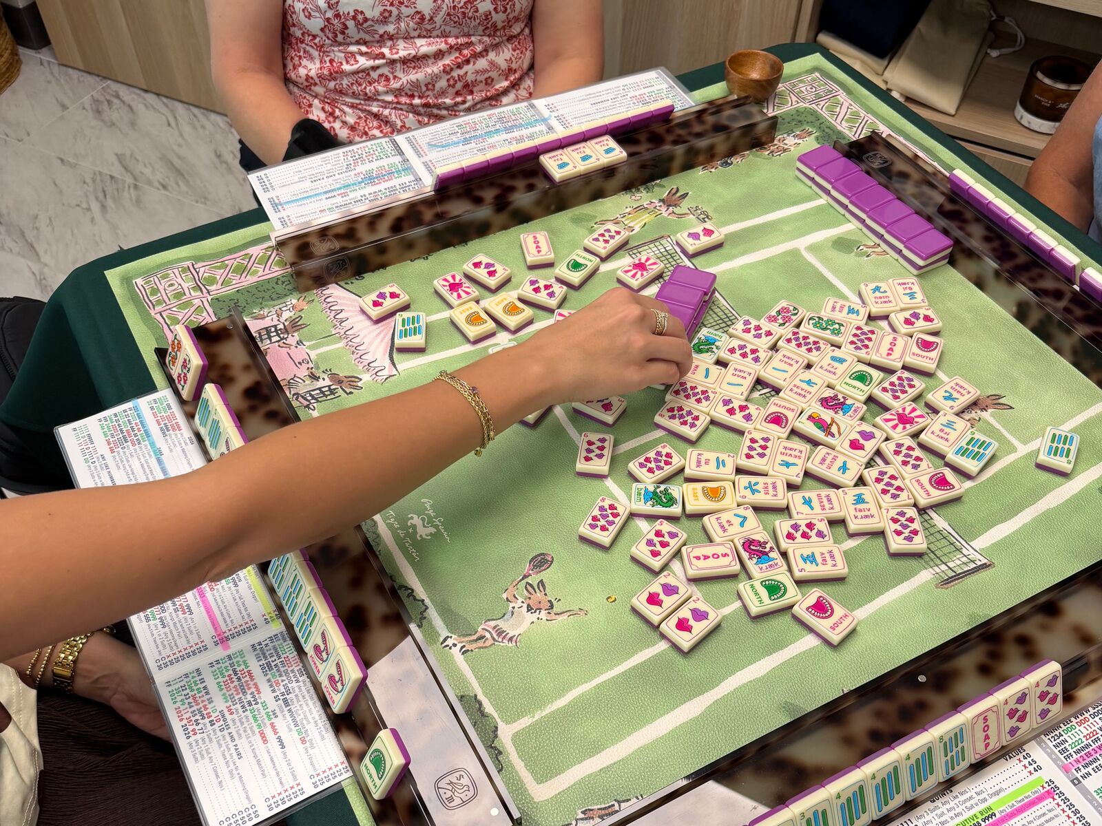 Hands reaching for tiles on a green pagoda-print Mahjong mat with racks and NMJL cards