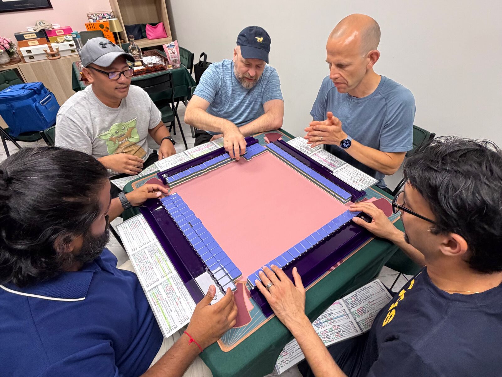 Four men seated around a pink Mahjong mat during Open Play, studying their racks and NMJL cards