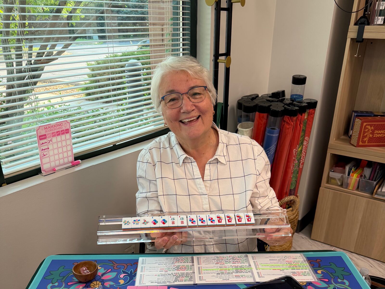 A smiling player holding up her rack of tiles above a blue and teal Mahjong mat