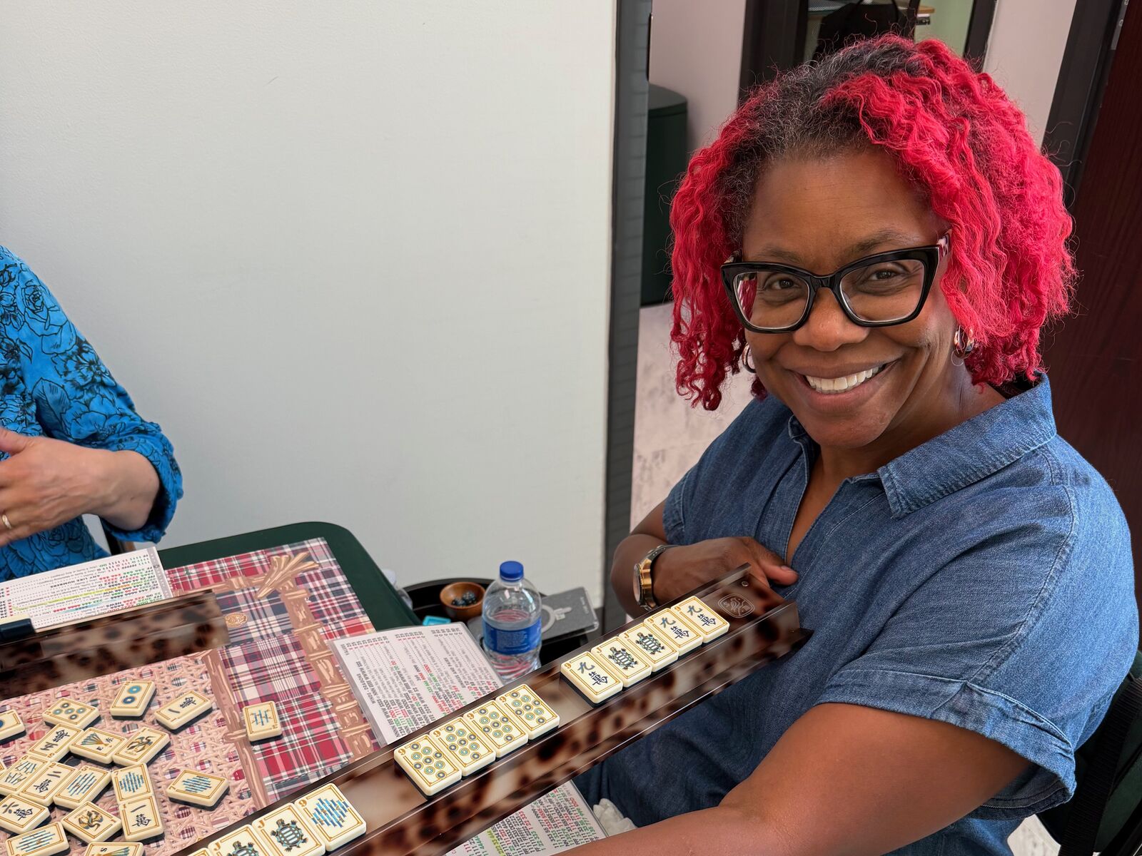 A smiling player showing her rack of tiles beside a plaid Mahjong mat