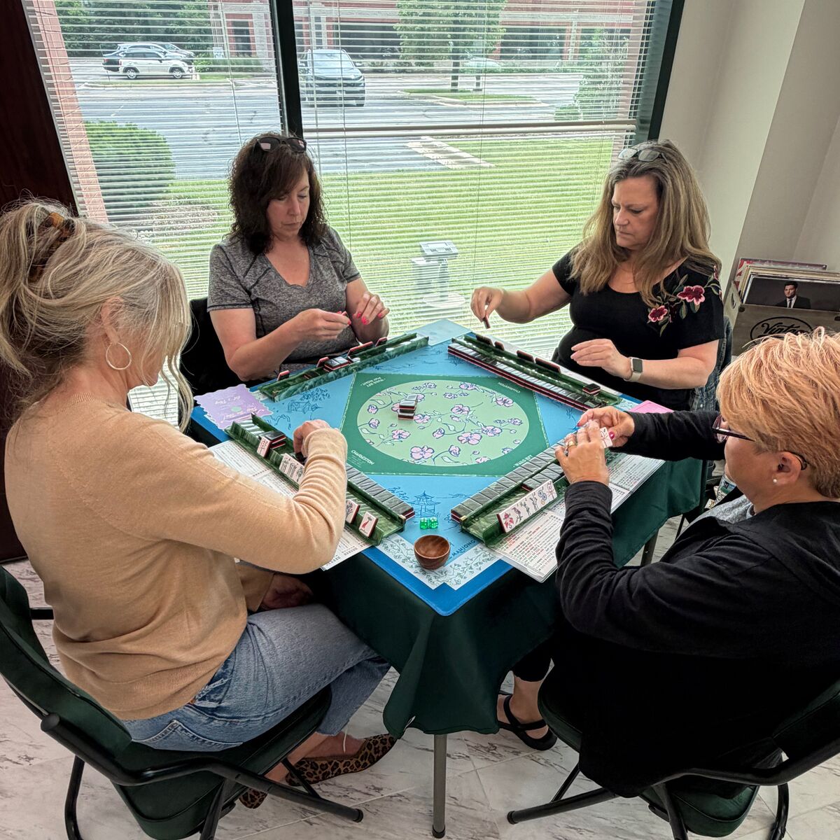 Four women playing American Mahjong around a blue and green table beside the windows at the Unique Mahjong Parlor